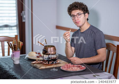 Young man sitting at a table enjoying burgundian fondue with boiling oil, skewering and eating meat, glass of red wine nearby in a cozy apartment dining scene Young man sitting at a table enjoying burgundian fondue with boiling oil, skewering and eating meat, glass of red wine nearby in a cozy apartment dining scene 140483977