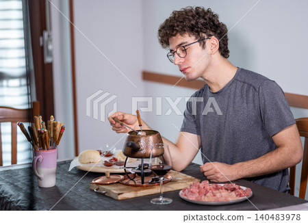 Young man enjoying a traditional fondue bourguignonne solo, cooking raw beef pieces in boiling oil with a fondue fork, surrounded by dipping sauces and a glass of red wine on a dark table 140483978