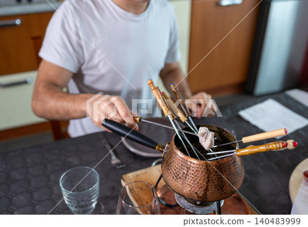 Man cooks beef pieces in a bubbling oil fondue pot at the table, enjoying a social, gourmet dining experience with friends or family in a cozy indoor setting 140483999