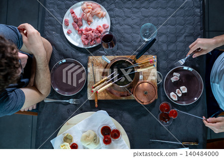 People are sitting at a table sharing a fondue bourguignonne meal, cooking various types of raw meat like beef and chicken in boiling oil, with different sauces and wine for a social dinner 140484000