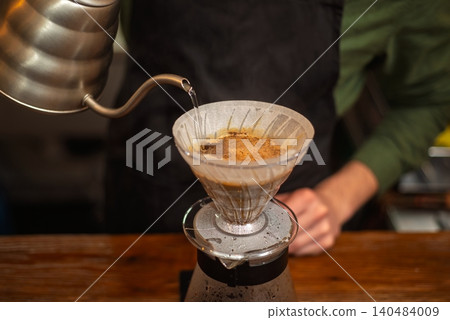 A close up of a barista pouring hot water from a gooseneck kettle into a coffee filter A close up of a barista pouring hot water from a gooseneck kettle into a coffee filter 140484009