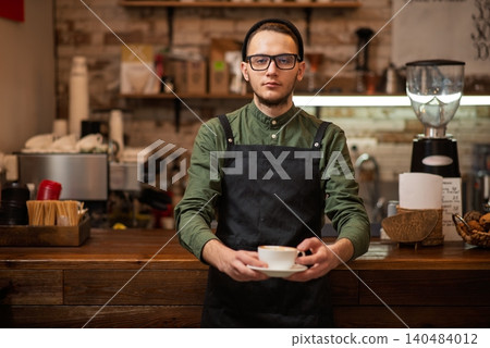 A professional male barista serving a fresh cup of coffee at a wooden counter 140484012