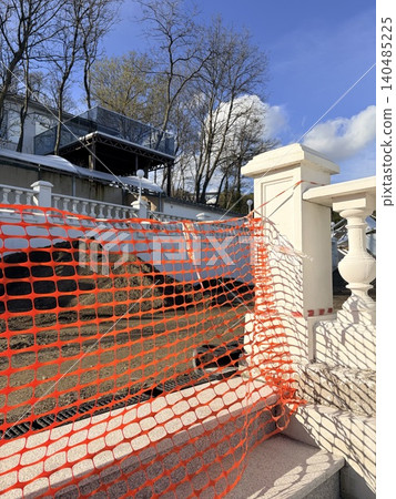 An orange plastic safety fence. a bright red protective mesh fence enclosing a construction site An orange plastic safety fence. a bright red protective mesh fence enclosing a construction site 140485225