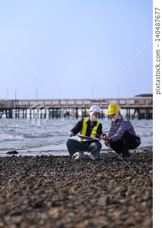 Two engineers stand on a rocky shoreline. They review data on tablets and devices. Pier into calm waters behind them 140487677
