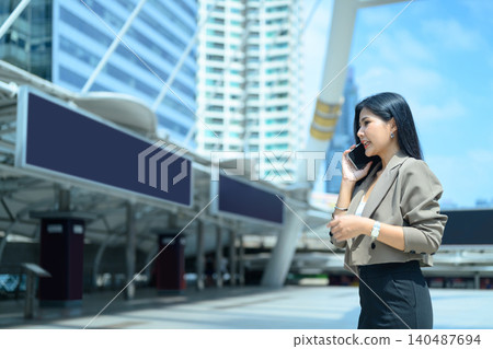 Smiling Asian business woman entrepreneur wearing suit talking with mobile phone standing in city metro Smiling Asian business woman entrepreneur wearing suit talking with mobile phone standing in city metro 140487694