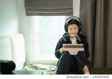 Happy Asian Student Girl Wearing Headphones Using Tablet on Bed in Dorm Room by Window Happy Asian Student Girl Wearing Headphones Using Tablet on Bed in Dorm Room by Window 140487695