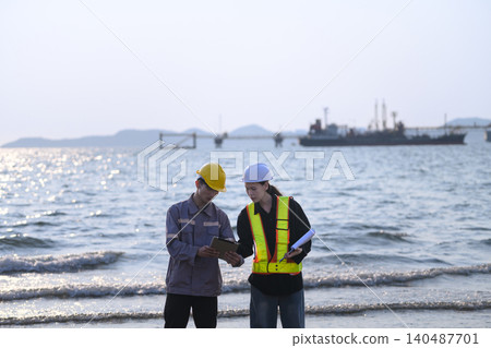 Two engineers stand on a rocky shoreline. They review data on tablets and devices. Pier into calm waters behind them Two engineers stand on a rocky shoreline. They review data on tablets and devices. Pier into calm waters behind them 140487701