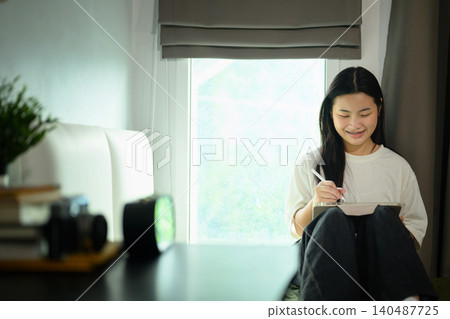 Young Asian Female Student Writing in Tablet on Bed in Cozy Dorm Room by Window 140487725