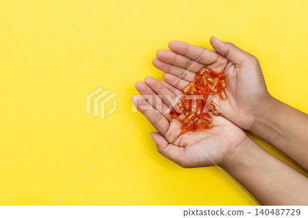 Hands cradle glowing orange capsules against bright yellow background. Soft lighting highlights each pill's glossy texture. Warm tone suggests health, wellness, or self-care Hands cradle glowing orange capsules against bright yellow background. Soft lighting highlights each pill's glossy texture. Warm tone suggests health, wellness, or self-care 140487729