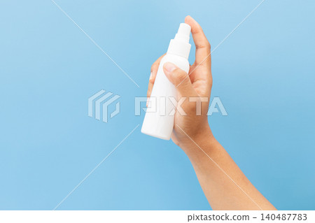Hand holds white spray bottle against soft blue background. Fingers grip bottle gently, showcasing clean, minimal design. Slight angle highlights spray nozzle and smooth surface 140487783