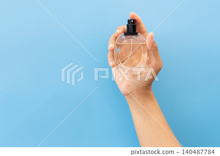 Hand holds clear perfume bottle against blue backdrop. Fingertips cradle glass, black nozzle visible. Simple, clean, minimalist aesthetic dominates frame. Ideal for beauty, lifestyle 140487784