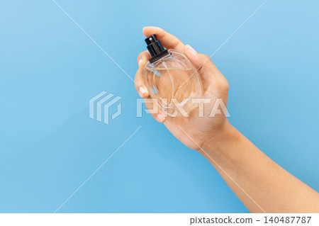 Hand holds clear perfume bottle against blue backdrop. Fingertips cradle glass, black nozzle visible. Simple, clean, minimalist aesthetic dominates frame. Ideal for beauty, lifestyle Hand holds clear perfume bottle against blue backdrop. Fingertips cradle glass, black nozzle visible. Simple, clean, minimalist aesthetic dominates frame. Ideal for beauty, lifestyle 140487787