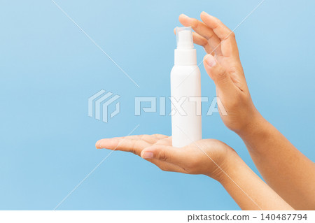 Hand holds white spray bottle against soft blue background. Fingers grip bottle gently, showcasing clean, minimal design. Slight angle highlights spray nozzle and smooth surface 140487794