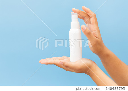 Hand holds white spray bottle against soft blue background. Fingers grip bottle gently, showcasing clean, minimal design. Slight angle highlights spray nozzle and smooth surface 140487795