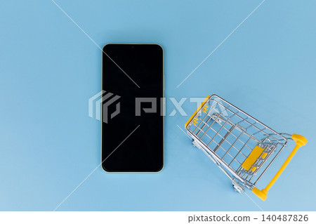 Minimalist scene with smartphone and shopping cart. Flat lay on solid blue background. Modern device beside miniature retail cart. Suggests online shopping or e-commerce concept 140487826