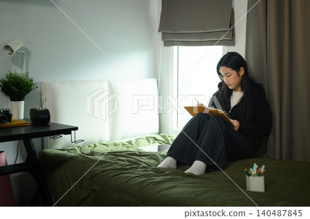 Young Asian Female Student Writing in Notebook on Bed in Cozy University Dorm Room by Window Young Asian Female Student Writing in Notebook on Bed in Cozy University Dorm Room by Window 140487845