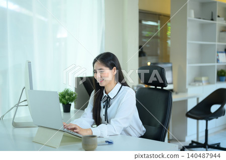 Cheerful young businesswoman working on laptop at modern office, professional woman using computer for remote work or online business in workspace 140487944