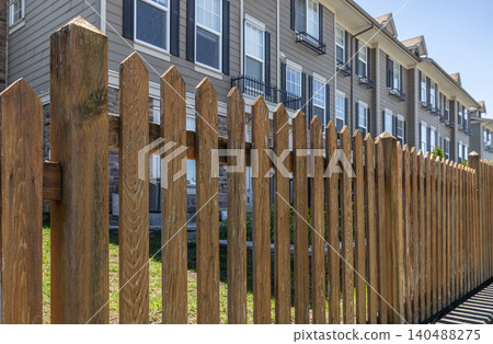 Wooden brown picket fence in residential neighborhood with green lawn in the garden. Townhouses with new wooden fence around the yard. Wooden fence with green lawn. Street photo, nobody Wooden brown picket fence in residential neighborhood with green lawn in the garden. Townhouses with new wooden fence around the yard. Wooden fence with green lawn. Street photo, nobody 140488275