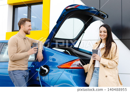 Young couple man and woman traveling by electric car having stop at charging station. Boyfriend plugging in cable to charge. Young couple man and woman traveling by electric car having stop at charging station. Boyfriend plugging in cable to charge. 140488955