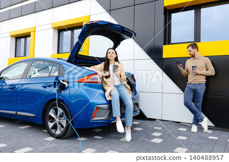 Young couple man and woman traveling together by new car having stop at charging station. Boyfriend holding cup drinking hot coffee talking to the phone and girlfriend looking aside and smiling Young couple man and woman traveling together by new car having stop at charging station. Boyfriend holding cup drinking hot coffee talking to the phone and girlfriend looking aside and smiling 140488957