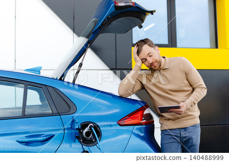 A young man is charging an electric car while standing at a station and holding a tablet A young man is charging an electric car while standing at a station and holding a tablet 140488999