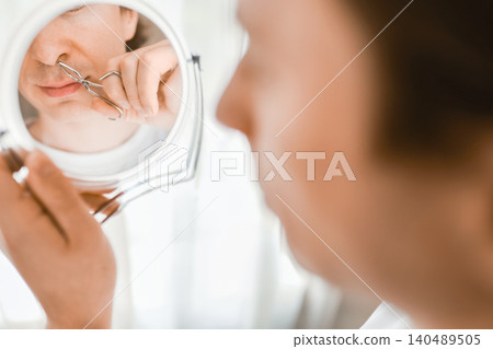 A man uses small tweezers for nose hair removal while looking in a mirror. Daily male hygiene, facial care, and grooming routine. A man uses small tweezers for nose hair removal while looking in a mirror. Daily male hygiene, facial care, and grooming routine. 140489505