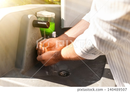 A man washing his hands under running water at an outdoor portable sink with soap. Essential hand hygiene, sanitation, and disease prevention at a campground or outdoor event. A man washing his hands under running water at an outdoor portable sink with soap. Essential hand hygiene, sanitation, and disease prevention at a campground or outdoor event. 140489507