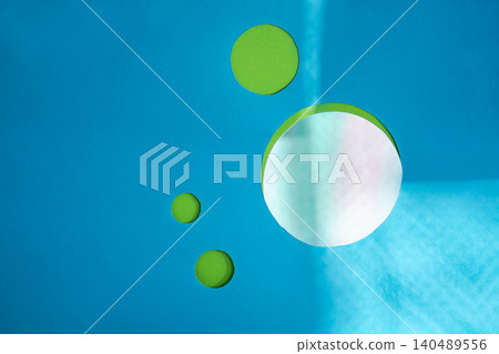 Green circles cut from paper are arranged on bright turquoise blue background in varying sizes. Large round shape features reflective, iridescent surface with soft light catching its edge. 140489556