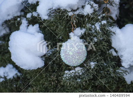White glitter christmas bauble hanging on snow covered garden shrub with colorful foliage and soft bokeh background. White iridescent glitter christmas bauble White glitter christmas bauble hanging on snow covered garden shrub with colorful foliage and soft bokeh background. White iridescent glitter christmas bauble 140489594