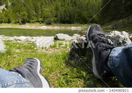 Person in hiking boots relaxing by a lake surrounded by forest and mountains, highlighting lakeside tourism and alpine recreation Person in hiking boots relaxing by a lake surrounded by forest and mountains, highlighting lakeside tourism and alpine recreation 140490921