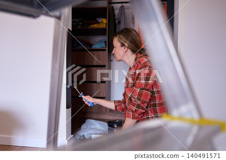 Woman applies dark paint to wall with roller, with ladder blurred in foreground Woman applies dark paint to wall with roller, with ladder blurred in foreground 140491571