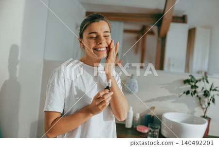 Happy young Caucasian woman applying cosmetic oil serum on face in bright sunny bathroom Happy young Caucasian woman applying cosmetic oil serum on face in bright sunny bathroom 140492201