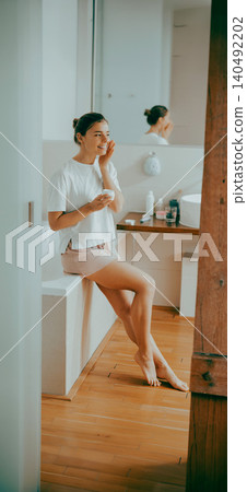 Caucasian young adult woman applying moisturizing cream to her face in a modern bathroom Caucasian young adult woman applying moisturizing cream to her face in a modern bathroom 140492202