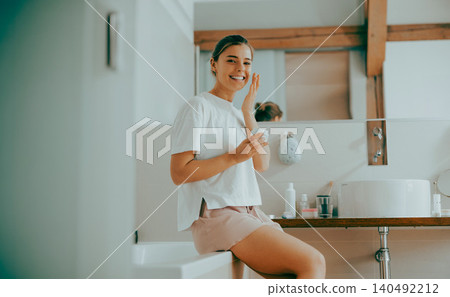 Smiling young Caucasian woman applying moisturizing face cream in bright modern bathroom Smiling young Caucasian woman applying moisturizing face cream in bright modern bathroom 140492212