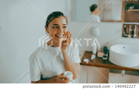 Happy Caucasian young adult woman applying moisturizing face cream in a bright bathroom Happy Caucasian young adult woman applying moisturizing face cream in a bright bathroom 140492227