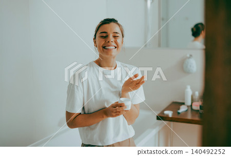 Happy young Caucasian woman applying skin cream while smiling in a bright bathroom Happy young Caucasian woman applying skin cream while smiling in a bright bathroom 140492252