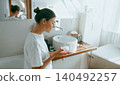 Happy young Caucasian woman holding skincare moisturizer jar in bright modern bathroom 140492257