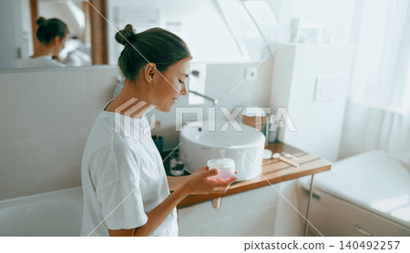 Happy young Caucasian woman holding skincare moisturizer jar in bright modern bathroom Happy young Caucasian woman holding skincare moisturizer jar in bright modern bathroom 140492257