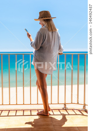 Woman in beach cover up and cowboy hat using smartphone on balcony with turquoise sea view, summer vacation remote Woman in beach cover up and cowboy hat using smartphone on balcony with turquoise sea view, summer vacation remote 140492487