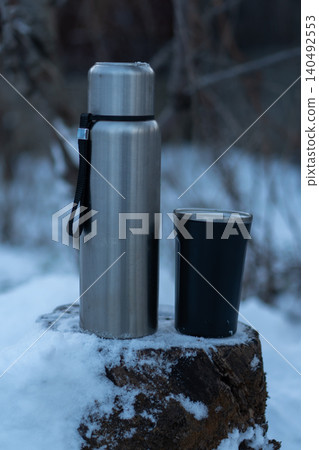 A thermos and a thermal mug on a snow-covered tree stump on a winter day. 140492553