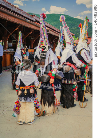 Kukeri group in masks and bell costumes gathers near mountain village during summer festival. Kukeri festival represents folklore, ritual dance, heritage tourism, carnival tradition Kukeri group in masks and bell costumes gathers near mountain village during summer festival. Kukeri festival represents folklore, ritual dance, heritage tourism, carnival tradition 140492990