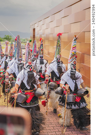 Kukeri performers in masks and bell costumes walk in village procession near building facade. Kukeri festival represents folklore, ritual dance, heritage tourism, carnival tradition Kukeri performers in masks and bell costumes walk in village procession near building facade. Kukeri festival represents folklore, ritual dance, heritage tourism, carnival tradition 140492994