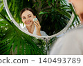 Smiling young Caucasian woman cleansing skin with facial device in mirror at bathroom 140493789