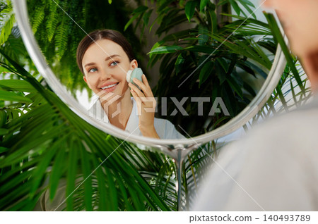 Smiling young Caucasian woman cleansing skin with facial device in mirror at bathroom Smiling young Caucasian woman cleansing skin with facial device in mirror at bathroom 140493789