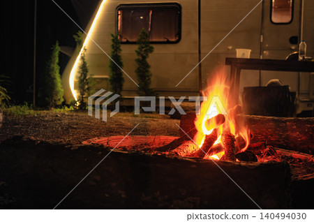 Glowing campfire surrounded by logs in front of a warmly lit camper, creating a cozy night camping vibe. 140494030