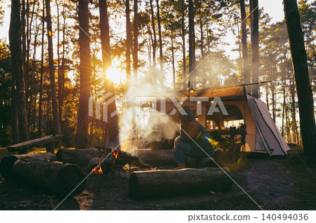 Camper sits by a crackling fire surrounded by logs in a serene forest, with sunlight streaming through the trees and a cozy tent in the background. Camper sits by a crackling fire surrounded by logs in a serene forest, with sunlight streaming through the trees and a cozy tent in the background. 140494036