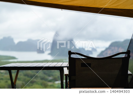 Cozy table and camping chair are set outside the tent, overlooking the sea and mist-covered mountains, amidst the serene and beautiful nature of Thailand. 140494303
