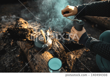 Serene forest camp showing a kettle, an enamel mug, and a person holding a coffee grinder on a log. Warm sunlight filters through the trees, creating a cozy and rustic vibe. Serene forest camp showing a kettle, an enamel mug, and a person holding a coffee grinder on a log. Warm sunlight filters through the trees, creating a cozy and rustic vibe. 140494308
