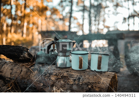 Cozy forest camp with a kettle, moka pot, and two enamel mugs on a log. Surrounded by sunlight, trees, and smoke from the fire, it radiates warmth and rustic simplicity. 140494311