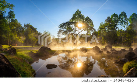 Serene hot spring during sunrise, with mist rising above reflective water, surrounded by rocks, trees, and lush greenery. The golden sunlight creates a tranquil and natural atmosphere. Serene hot spring during sunrise, with mist rising above reflective water, surrounded by rocks, trees, and lush greenery. The golden sunlight creates a tranquil and natural atmosphere. 140494420